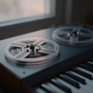 Close-up of a vintage analog synthesizer tape reel spinning slowly, desaturated charcoal tones, soft twilight light, 35mm film photography