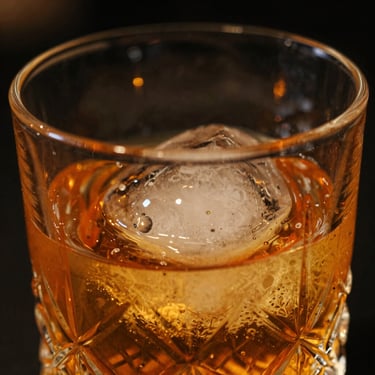 Macro close-up of a crystal cocktail glass with amber glowing liquid, ice sphere reflecting warm low-key lounge lights, dark background, cinematic depth of field.
