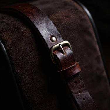 Dramatic low-key studio detail shot of a leather strap with metal hardware, chiaroscuro lighting, deep shadows, rich dark tones.