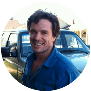 Smiling man in a blue work shirt standing in front of a silver pickup truck outdoors.