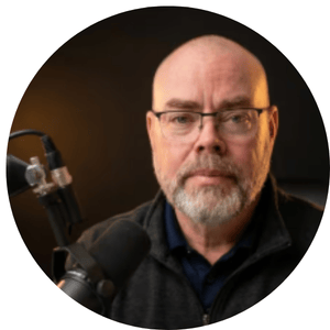 Professional male podcaster with glasses and a grey beard speaking into a studio microphone.