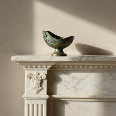 Tight tactile close-up of a premium marble fireplace mantel with classical carvings, holding a single bronze sculptural object. Warm afternoon light casting subtle, elegant shadows on the limestone wall behind.