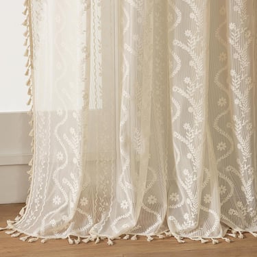 Elegant cream floral embroidered lace curtains with bohemian fringe tassels on a wooden floor.