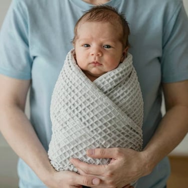 Close-up of gentle hands holding a newborn wrapped in a soft organic cotton waffle blanket, diffused daylight