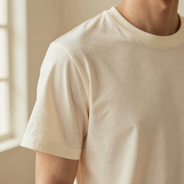 Close-up of a model wearing a heavyweight cream T-shirt, highlighting the structured shoulder seam and thick fabric drape, warm studio lighting
