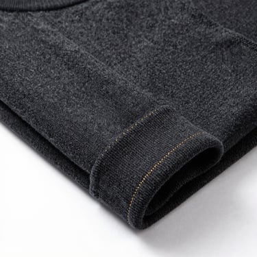 High-end studio product shot of a folded dark charcoal French terry crewneck sweater, showing detailed gold stitching on the cuff, clean architectural presentation.