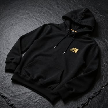 High-end studio product shot of a structured matte black hoodie with clean gold embroidery on the chest, flat lay on a dark slate surface, dramatic low-contrast lighting.