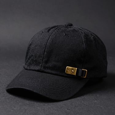 High-end studio product shot of a structured low-profile black cotton cap with a brass buckle, set against a dark grey background, soft side lighting highlighting the texture.