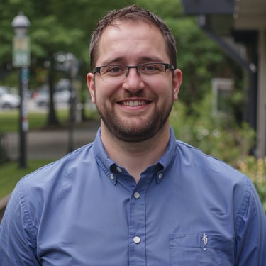 A smiling man with glasses and a beard wearing a blue button-down shirt outdoors.