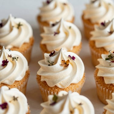 Editorial eye-level shot of six silky Swiss meringue cupcakes, piped with immaculate precision, decorated with hand-pressed botanicals.
