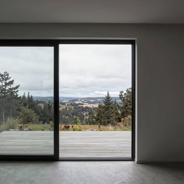 Architectural interior view of a minimal villa showing a flush threshold sliding glass door opening directly onto a wooden deck, overcast daylight