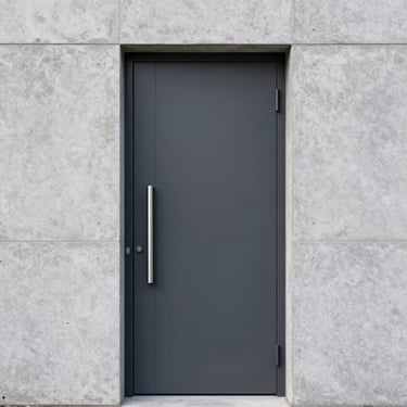 A tall, minimalist dark charcoal entrance door with a long vertical brushed steel pull handle, set in a light grey concrete facade