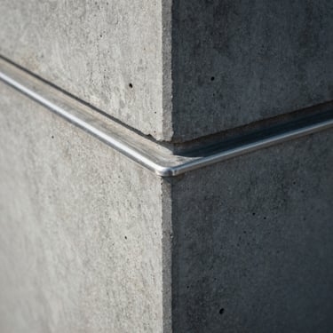 Tight macro crop of architectural concrete lines meeting a brushed steel edge, cool diffused daylight, high-contrast shadows.