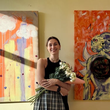 an artist in exhibition standing next to her paintings holding flowers