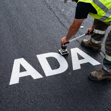 Overhead shot of an ADA stencil being laid down on fresh asphalt by a crew member in safety gear.