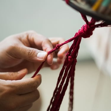 Manos tejiendo macramé artesanal con hilo rojo, proceso de creación hecho a mano.