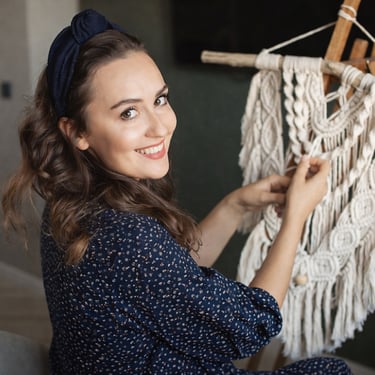 Artesana trabajando un tapiz de macramé hecho a mano, sonriendo mientras ajusta los nudos en un soporte de madera.