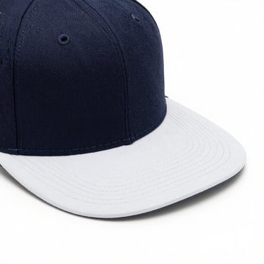 Structured snapback cap in navy with a clean white brim, 45-degree angle on white surface, studio lighting, sharp stitching detail, no people