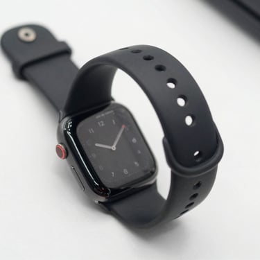 45-degree product shot of a smartwatch with a black sport band on a clean white surface, watch face displaying time, sharp studio lighting