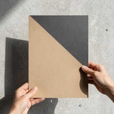 A hand holding a heavy, textured paper swatch against a clean concrete background, sharp sunlight, architectural focus