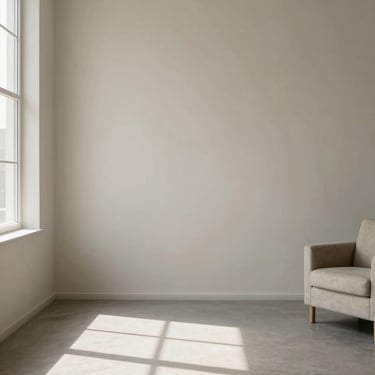 Wide architectural interior of a minimalist therapy room, natural north-facing window light falling across a pale plaster wall to the left third, an empty armchair partially visible at the right edge, warm gray and off-white tones, no people, no decor, geometric shadow lines from window frame crossing the floor