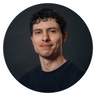 Professional headshot of a smiling young man with curly dark hair and a navy blue t-shirt.