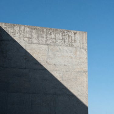 A minimalist concrete architectural corner meeting a clear blue sky, harsh high-contrast natural light casting a razor-sharp diagonal shadow.