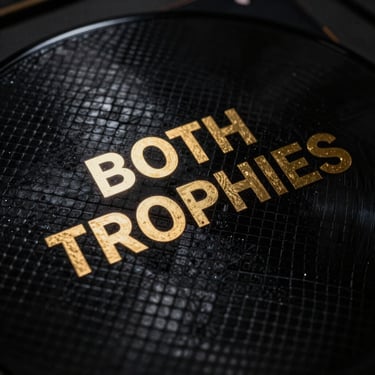 Extreme close-up of a luxury vinyl record sleeve with gold foil lettering reading BOTH TROPHIES, resting on textured black snakeskin under high-contrast dramatic studio lighting.