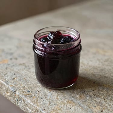 Elegant glass jar of dark berry jam on a rustic stone counter, soft golden light catching the rich texture