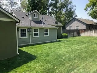 Side view of an olive student house near BSU with a green lawn, white-trimmed windows, and a backyard privacy fence.