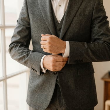 Man in a perfectly tailored charcoal grey suit adjusting his cufflinks, warm natural light, square aspect