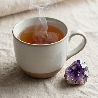 A ceramic mug of warm herbal tea with steam rising, next to a polished amethyst crystal on a soft linen cloth, diffused light.