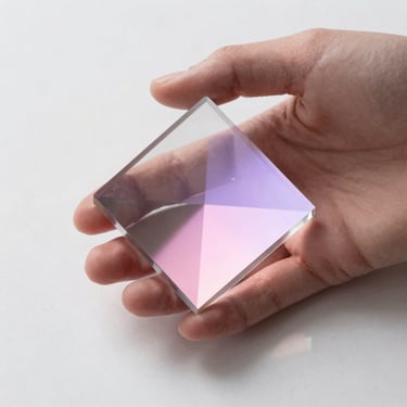 A delicate hand holding a glass prism reflecting pastel pink and lavender light onto a clean white surface, soft focus.