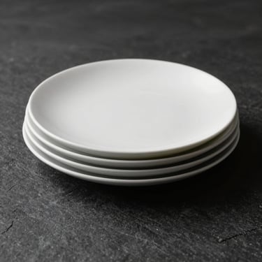 Minimalist composition of fine matte white ceramic plates stacked on a dark stone surface, deep shadows