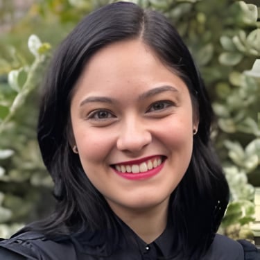 Smiling woman with dark hair and red lipstick posing outdoors against green foliage.