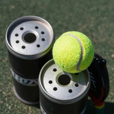 Overhead close-up of three padel ball cans stacked on a court surface under daylight, the yellow-green felt of one open can visible