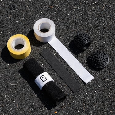 Overhead flat lay of padel accessories — grip tape rolls, overgrip strips, wristbands, and a vibration dampener on a dark court surface under sharp daylight