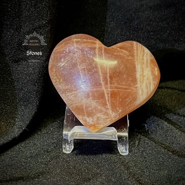 A sunstone/moonstone heart crystal displayed on a clear stand against a black fabric background.