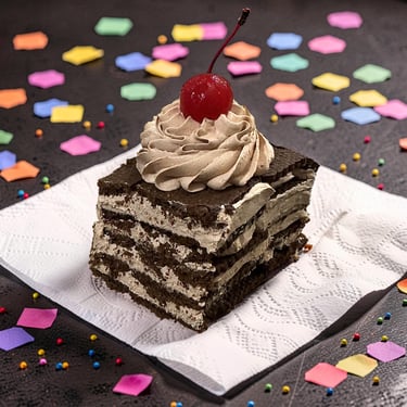 A chocolate layered cake slice with whipped cream frosting and a cherry on top with colorful confetti.