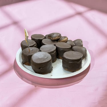 Un plato blanco lleno de malvaviscos gourmet cubiertos de chocolate sobre una superficie rosa con so
