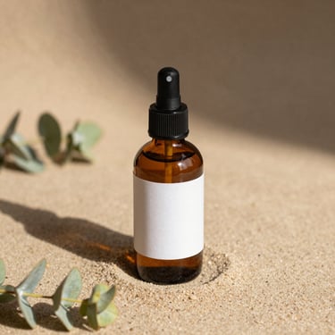 A clean amber glass spray bottle with a minimalist label, resting on a flat warm sand surface with soft shadows of eucalyptus leaves.