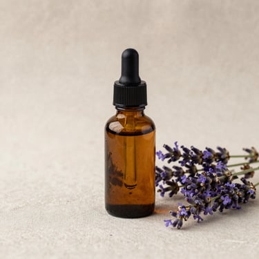 An amber glass dropper bottle sitting next to fresh lavender blossoms on a soft beige linen surface, gentle side light, minimalist aesthetic.
