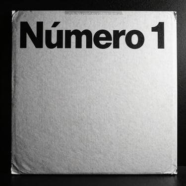 High-contrast black and white photo of the original 1990 'Número 1' vinyl sleeve, harsh flash, deep shadows, showing the minimalist typography on textured cardboard.