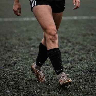 Tight crop on a female soccer player's muddy cleats and lower legs in motion on turf, high-contrast desaturated tones, 35mm.
