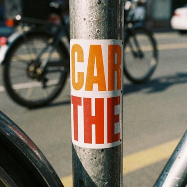 Two high-contrast vinyl stickers with bold typography, one orange and one red, stuck to a scuffed metal bike frame, motion-blurred street background, 35mm film