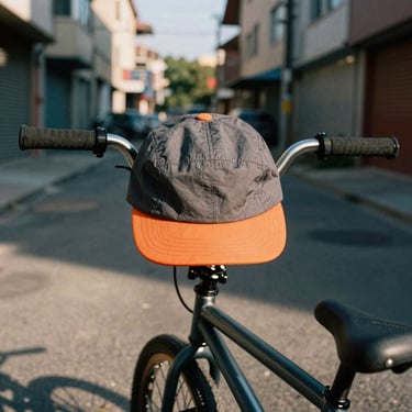 A classic five-panel cycling cap in dark grey with an orange brim, hanging from a set of BMX handlebars in an urban alley, high-contrast street tones, 35mm film