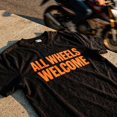 A close-up of a high-contrast black t-shirt with a bold orange 'ALL WHEELS WELCOME' print, resting on a concrete ledge, warm golden-hour sunlight, gritty 35mm film style