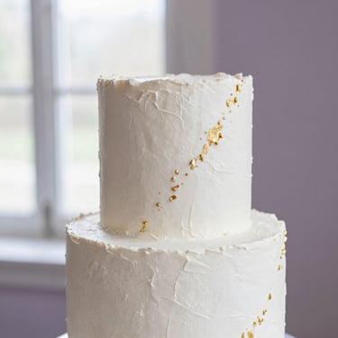A minimalist wedding cake with clean white textured buttercream and subtle gold leaf accents, elegant styling.