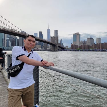 A man posing by the East River with the Brooklyn Bridge and New York City skyline in the background.