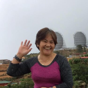 Smiling woman waving in front of large satellite dishes on a hillside landscape.